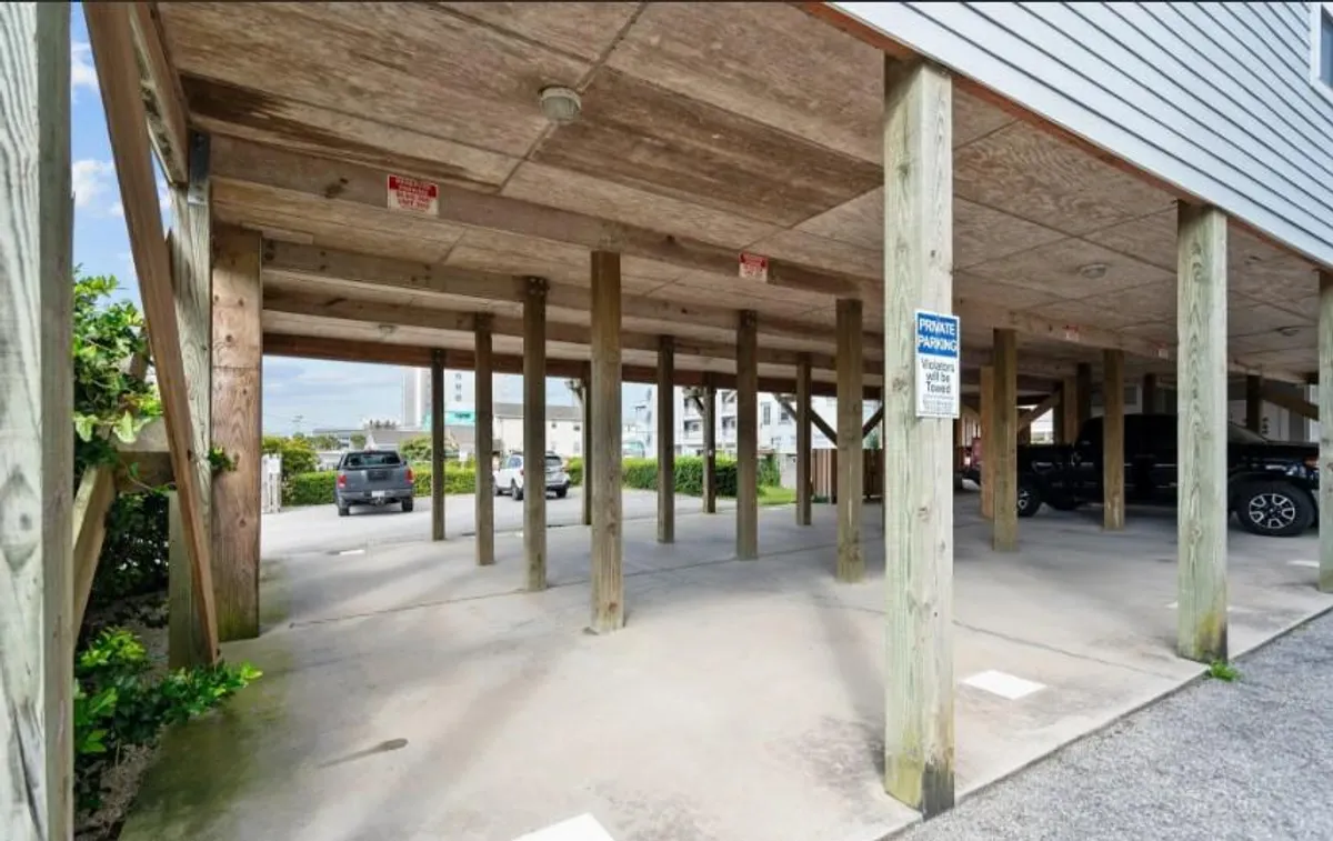 A practical ground-level area beneath the condo, featuring wooden support beams, parking signs, concrete flooring, and surrounding green foliage. The scene suggests easy access to both the beach and outdoor equipment, with open air and a connection to the outside world.