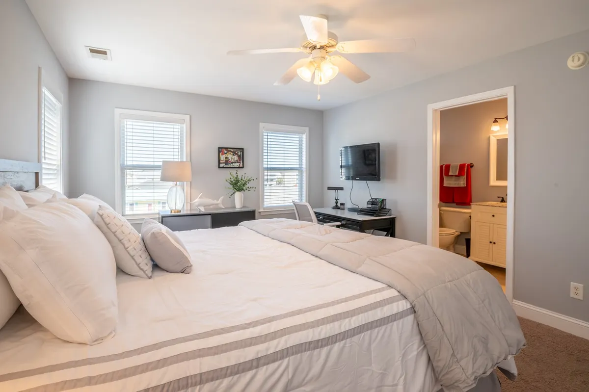 A cozy bedroom with gray and white bedding, large windows letting in daylight, a wall-mounted TV, and a small plant on the desk. The room feels inviting and perfect for restful nights.