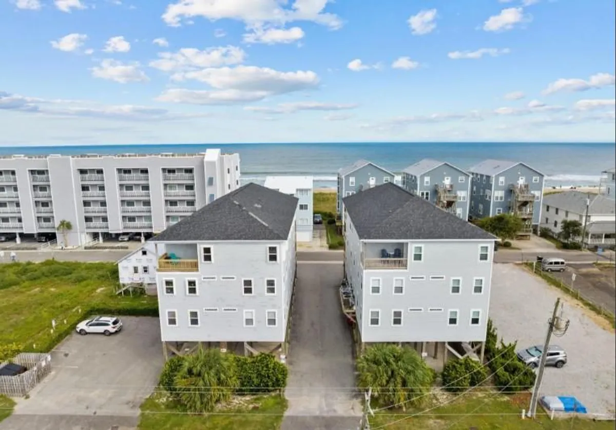 This image showcases a beachfront view with two main buildings, surrounded by lush greenery and several parked cars. The inviting and tranquil mood is highlighted by visible beach activities, families gathering near the shore, and children playing in the sand. The proximity to the ocean and openness of the area emphasize the ease of beach access for guests.