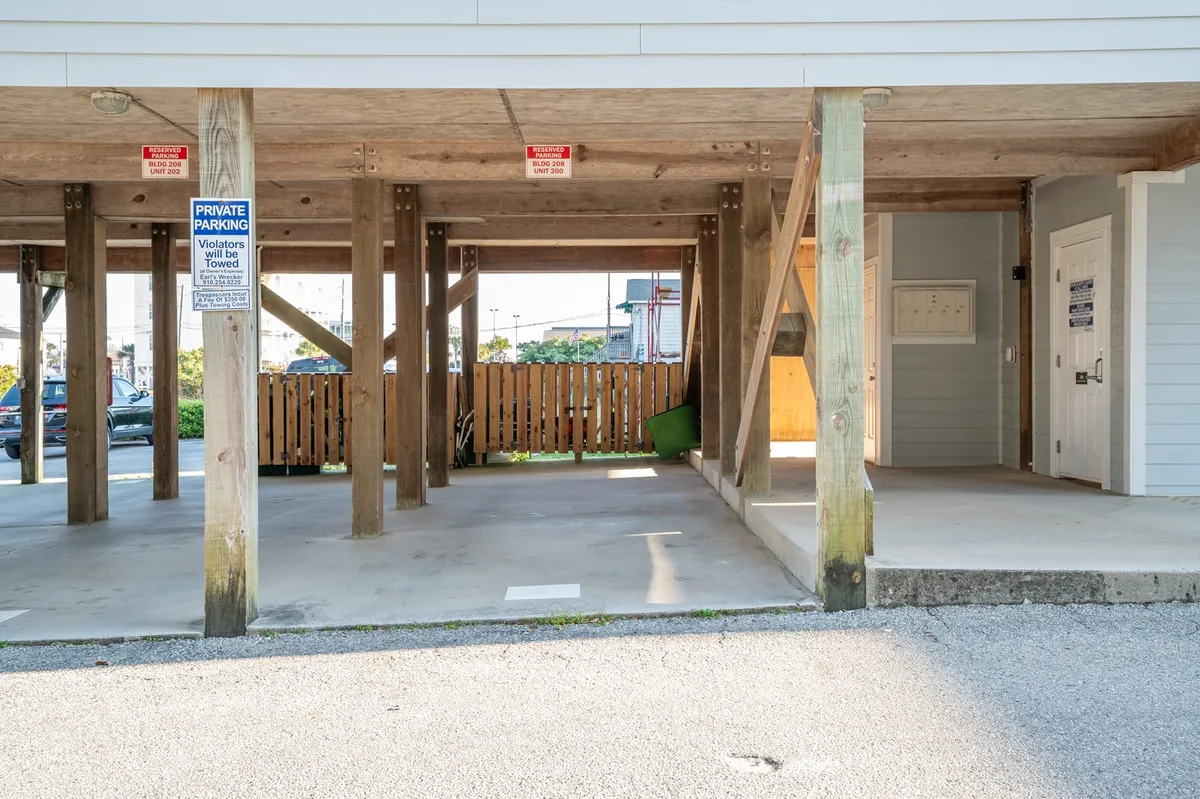 An exterior ground-level view beneath the building, showing wooden support beams, parking signage, and an open shaded space. The area appears inviting for gatherings or as a convenient spot before heading out to the nearby boardwalk, restaurants, or nightlife.