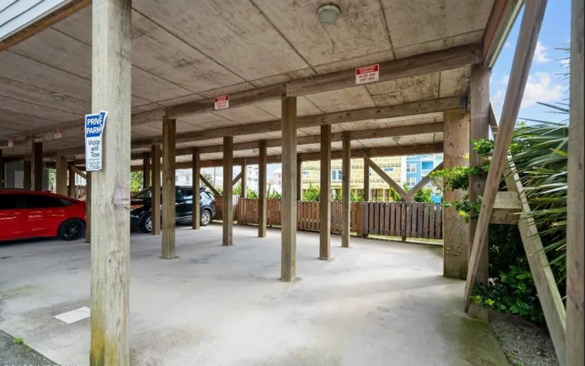 A view of the covered parking area beneath the building, with vehicles parked and visible support beams and signage. The space is utilitarian, offering shelter and convenient access for residents.